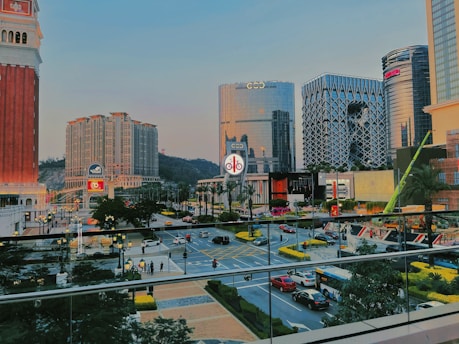 A cityscape with modern high-rise buildings featuring unique architectural designs, surrounded by lush greenery. The area is bustling with traffic, including cars and buses, on a multi-lane road with decorative street lamps. A pedestrian walkway is visible with a few people walking along it.