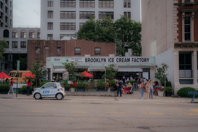 A bustling street scene featuring the Brooklyn Ice Cream Factory, surrounded by people enjoying the outdoor seating under red umbrellas. A small NYPD car is parked in front, and tall buildings loom in the background. Lush greenery adds vibrancy to the urban setting, and people are casually dressed, indicating a relaxed atmosphere.