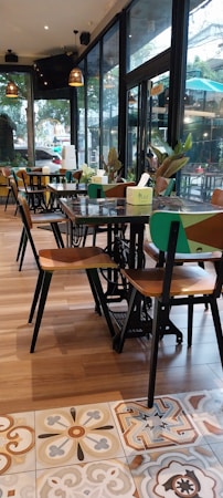 A cozy, modern caf&eacute; interior with wooden flooring and patterned tiles. The caf&eacute; features wooden tables and chairs, with some tables adorned with green plants and small paper menus. Large windows allow natural light to flood the space, and there are hanging pendant lights providing a warm ambiance. The view through the windows shows greenery and a hint of urban environment.