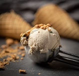 A creamy scoop of ice cream topped with small crunchy pieces is placed in a metal ice cream scoop. In the background, there are waffle cones slightly out of focus. Crumbs are scattered on a dark surface, adding texture to the scene.