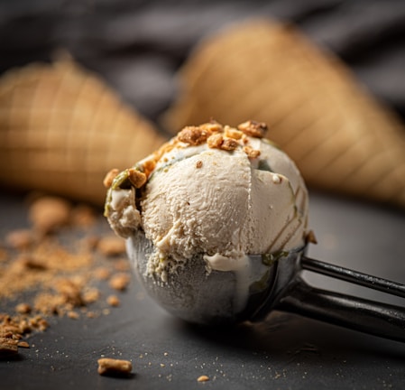 A creamy scoop of ice cream topped with small crunchy pieces is placed in a metal ice cream scoop. In the background, there are waffle cones slightly out of focus. Crumbs are scattered on a dark surface, adding texture to the scene.