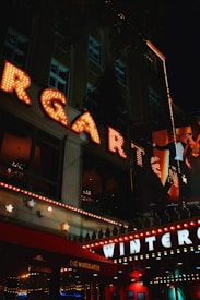 Bright neon lights illuminate a nighttime urban scene, prominently displaying large letters spelling out 'WINTERGARTEN'. The entrance is adorned with warm, glowing bulbs and decorative stars, creating a festive atmosphere. A café named 'Café Wintergarten' is visible, with people in formal attire adding a sense of elegance.