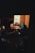 A cozy, dimly lit room with vintage furniture, including a wooden round table surrounded by armchairs. A warm light from a table lamp casts a glow over the scene. The decor features patterned cushions and a relaxed, intimate atmosphere.