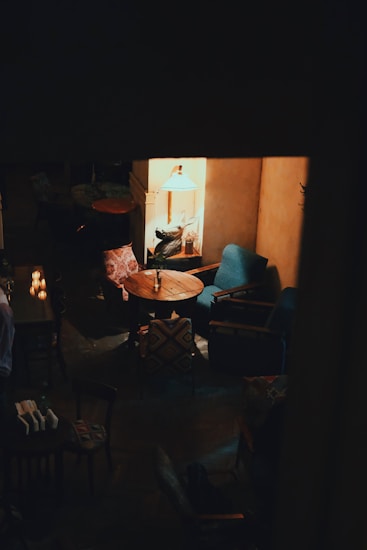A cozy, dimly lit room with vintage furniture, including a wooden round table surrounded by armchairs. A warm light from a table lamp casts a glow over the scene. The decor features patterned cushions and a relaxed, intimate atmosphere.