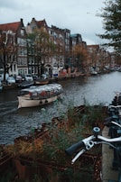 A canal in an urban setting lined with historic buildings featuring traditional architecture. A glass-roofed boat is navigating the waterway, with bicycles parked along the sidewalk. Trees with autumn foliage add a touch of color to the scene.