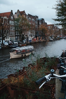 A canal in an urban setting lined with historic buildings featuring traditional architecture. A glass-roofed boat is navigating the waterway, with bicycles parked along the sidewalk. Trees with autumn foliage add a touch of color to the scene.