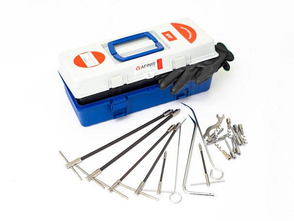 A blue and white toolbox containing various precision tools laid out neatly beside it. The tools appear to be specialized dental or surgical instruments made of metal. There are also black gloves placed on top of the toolbox.