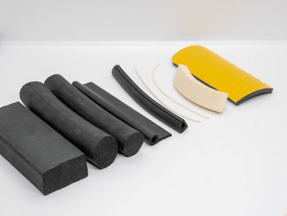 Various pieces of foam and rubber insulation materials are neatly arranged on a white surface. The objects vary in shape from rectangular blocks to curved and tubular forms. They display a combination of colors including black, white, and yellow.