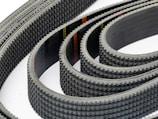 Multiple toothed belts are arranged in a curved, overlapping pattern. The belts display an industrial, textured surface with visible grooves and a dark color palette. Their consistent alignment and repetition create a sense of precision and complexity.