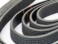 Multiple toothed belts are arranged in a curved, overlapping pattern. The belts display an industrial, textured surface with visible grooves and a dark color palette. Their consistent alignment and repetition create a sense of precision and complexity.