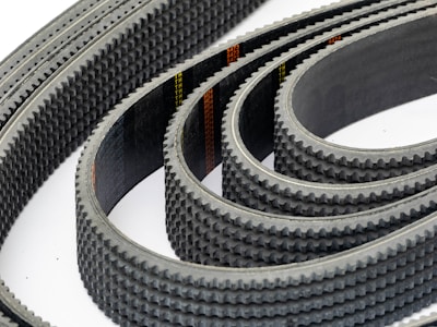 Multiple toothed belts are arranged in a curved, overlapping pattern. The belts display an industrial, textured surface with visible grooves and a dark color palette. Their consistent alignment and repetition create a sense of precision and complexity.