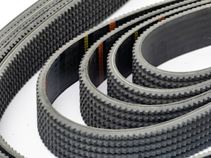 Multiple toothed belts are arranged in a curved, overlapping pattern. The belts display an industrial, textured surface with visible grooves and a dark color palette. Their consistent alignment and repetition create a sense of precision and complexity.