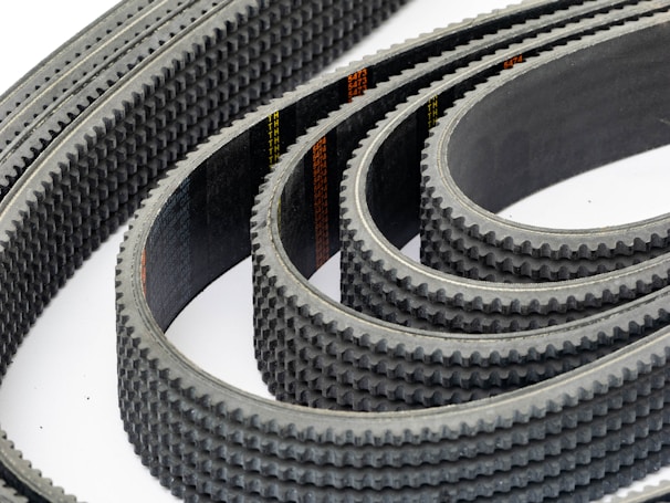 Multiple toothed belts are arranged in a curved, overlapping pattern. The belts display an industrial, textured surface with visible grooves and a dark color palette. Their consistent alignment and repetition create a sense of precision and complexity.