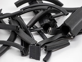 A collection of various black, cylindrical and rectangular rubber pieces with textured surfaces, some of which are partially open revealing metallic clips or inner components, arranged randomly on a flat, light-colored surface.
