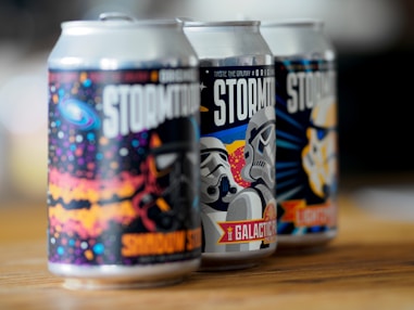 Three creatively designed beverage cans are placed in a row on a wooden surface. Each can features colorful and stylized artwork of space-themed imagery and stormtrooper-like helmets. The cans have strong contrasts and vibrant colors, offering a playful and sci-fi inspired aesthetic.