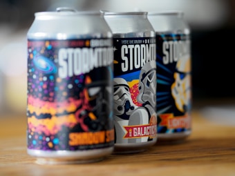 Three creatively designed beverage cans are placed in a row on a wooden surface. Each can features colorful and stylized artwork of space-themed imagery and stormtrooper-like helmets. The cans have strong contrasts and vibrant colors, offering a playful and sci-fi inspired aesthetic.