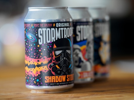 A close-up image of three beverage cans with sci-fi themed designs. The front can prominently displays a design with a helmeted figure, colorful stars, and cosmic graphics. The text on the can includes phrases like 'Stormtrooper' and 'Shadow Stout', along with the phrase 'Taste the Galaxy.' The other two cans in the background also have similar sci-fi-themed designs.