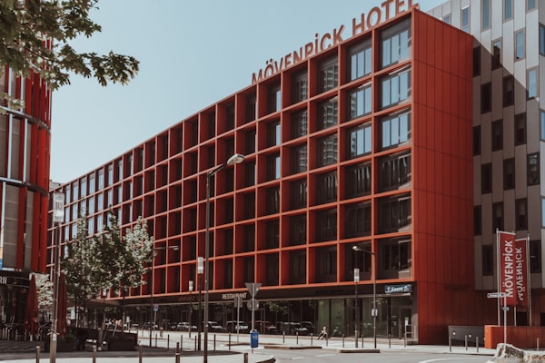 A modern hotel building with a red facade, featuring multiple windows and clean, geometric lines. The signage reads 'Mövenpick Hotel' and is prominently displayed at the top. The street is lined with small trees, streetlamps, and a few pedestrians. The sky is clear and blue, casting shadows on the building.
