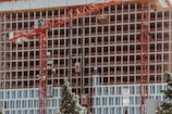A high-rise building under construction features multiple floors with red cranes positioned at the front. The structure displays a grid-like pattern with unfinished openings. Surrounding the site, green trees contrast with the industrial setting.