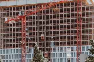 A high-rise building under construction features multiple floors with red cranes positioned at the front. The structure displays a grid-like pattern with unfinished openings. Surrounding the site, green trees contrast with the industrial setting.