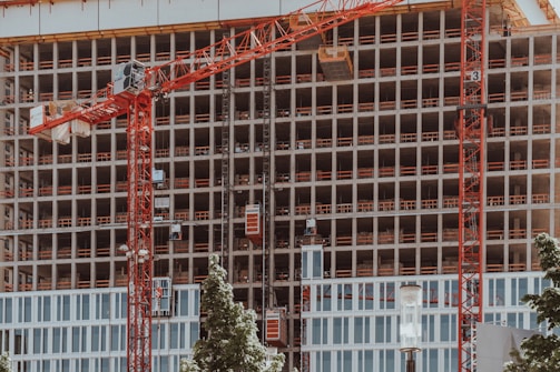 A high-rise building under construction features multiple floors with red cranes positioned at the front. The structure displays a grid-like pattern with unfinished openings. Surrounding the site, green trees contrast with the industrial setting.