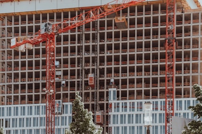A high-rise building under construction features multiple floors with red cranes positioned at the front. The structure displays a grid-like pattern with unfinished openings. Surrounding the site, green trees contrast with the industrial setting.