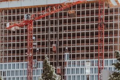 A high-rise building under construction features multiple floors with red cranes positioned at the front. The structure displays a grid-like pattern with unfinished openings. Surrounding the site, green trees contrast with the industrial setting.