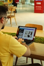 A person wearing a yellow shirt and a smartwatch is sitting at a wooden table in a café or workspace. They are using a laptop with graphic design software open. There is a patch of greenery on the table, and in the background, there are chairs and a person standing with a bag. The atmosphere is modern and casual.