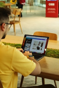 A person wearing a yellow shirt and a smartwatch is sitting at a wooden table in a café or workspace. They are using a laptop with graphic design software open. There is a patch of greenery on the table, and in the background, there are chairs and a person standing with a bag. The atmosphere is modern and casual.