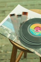A stack of vinyl records and film negatives are placed on top of newspapers, all resting on a wooden stool. The vinyl records feature a colorful label, while the film negatives have a classic brown tint. The background shows a blurred surface, possibly a textured floor.