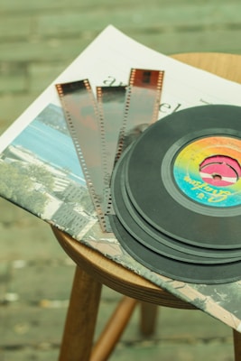 A stack of vinyl records and film negatives are placed on top of newspapers, all resting on a wooden stool. The vinyl records feature a colorful label, while the film negatives have a classic brown tint. The background shows a blurred surface, possibly a textured floor.