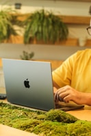 A person is working on a silver laptop with a distinctive logo, seated at a desk adorned with green moss. Decorative plants are visible in the background, conveying a natural and eco-friendly workspace atmosphere.