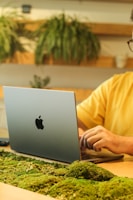 A person is working on a silver laptop with a distinctive logo, seated at a desk adorned with green moss. Decorative plants are visible in the background, conveying a natural and eco-friendly workspace atmosphere.