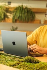 A person is working on a silver laptop with a distinctive logo, seated at a desk adorned with green moss. Decorative plants are visible in the background, conveying a natural and eco-friendly workspace atmosphere.