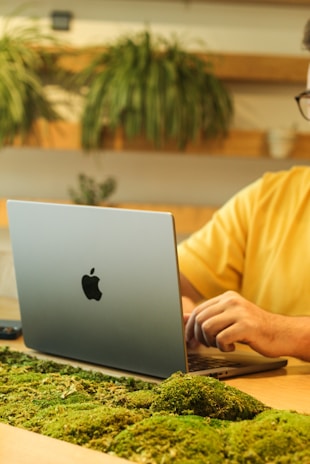 A person is working on a silver laptop with a distinctive logo, seated at a desk adorned with green moss. Decorative plants are visible in the background, conveying a natural and eco-friendly workspace atmosphere.