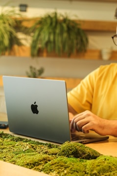 A person is working on a silver laptop with a distinctive logo, seated at a desk adorned with green moss. Decorative plants are visible in the background, conveying a natural and eco-friendly workspace atmosphere.