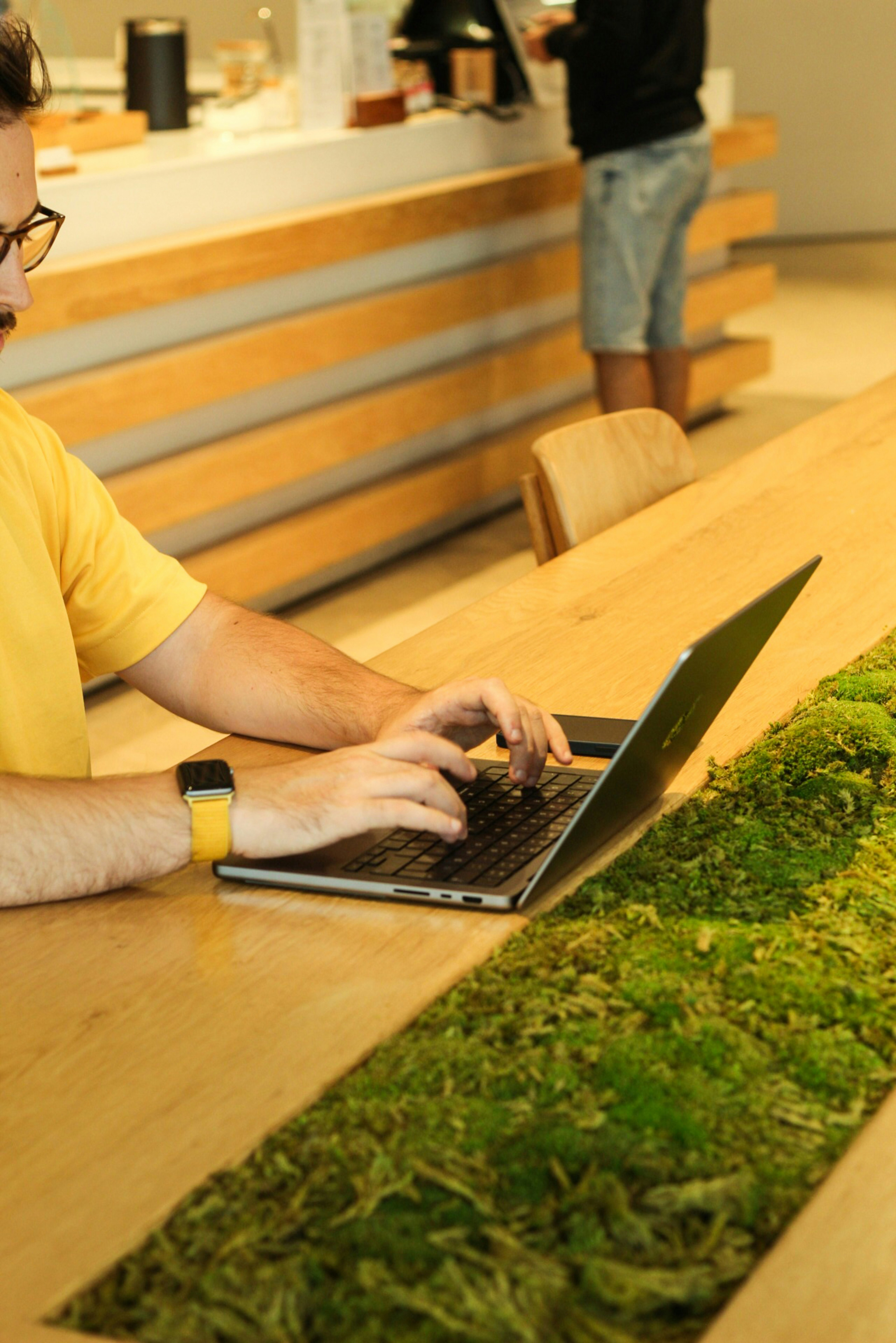 Man in a yellow shirt typing on a laptop at a moss table.