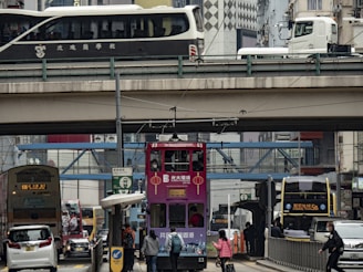 A bustling urban scene features a pink double-decker tram in the foreground, surrounded by various vehicles including buses and cars. Elevated highways are visible above, with buses and trucks traveling on them. Numerous pedestrians are visible, some waiting at a bus stop and others crossing the street. The cityscape is filled with tall buildings displaying advertising signs and architectural details.