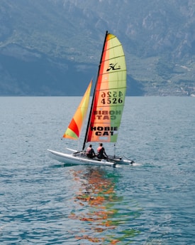 A catamaran with colorful sails is sailing on a body of water with two people on board. The water is a vivid blue-green, reflecting the colors of the sails. In the background, there are large rocky mountains partially covered with green foliage.