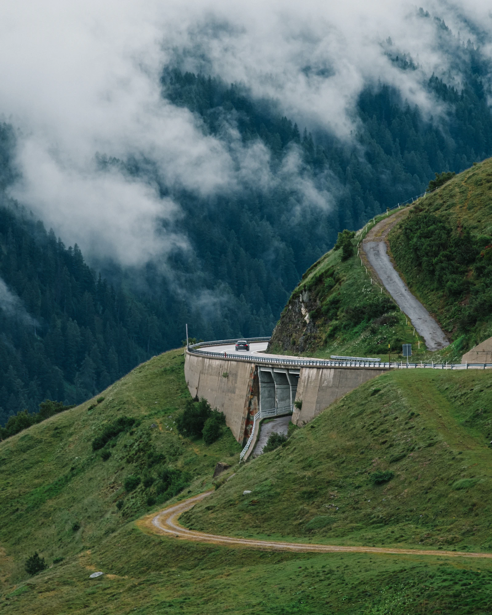 A Swiss road climbs beneath a bridge into low mountain cloud