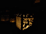 A lantern with intricate cut-out designs casting a warm glow in the dark. The designs feature a deer and foliage against a honeycomb pattern. The light creates subtle patterns and shadows around the lantern.
