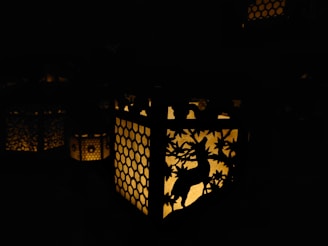 A lantern with intricate cut-out designs casting a warm glow in the dark. The designs feature a deer and foliage against a honeycomb pattern. The light creates subtle patterns and shadows around the lantern.