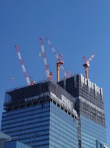 A high-rise building under construction with several red and white cranes at the top. The building is partially covered with black netting, suggesting ongoing construction work. The facade is composed of reflective glass windows, and the sky above is bright and clear.