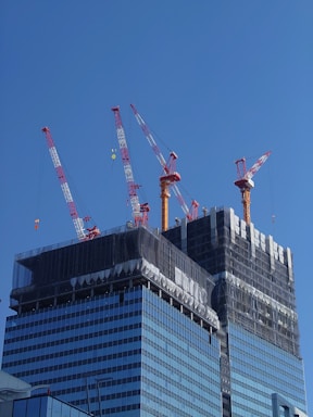 A high-rise building under construction with several red and white cranes at the top. The building is partially covered with black netting, suggesting ongoing construction work. The facade is composed of reflective glass windows, and the sky above is bright and clear.
