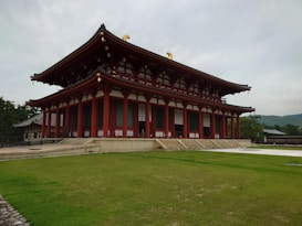 A traditional Japanese building with a striking red and white color scheme stands amidst a well-maintained lawn. The structure features intricate architectural details, including ornate roof tiles and decorative ridges. The sky above is overcast, creating a subdued atmosphere, while trees and hills are visible in the background.