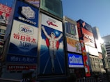 A vibrant urban scene featuring large, colorful billboards in a bustling cityscape. The central focus is a giant advertisement depicting a running man, surrounded by other illuminated signs showcasing various brands and products. The sky is clear, and the light suggests it is late afternoon.
