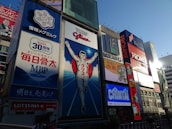 A vibrant urban scene featuring large, colorful billboards in a bustling cityscape. The central focus is a giant advertisement depicting a running man, surrounded by other illuminated signs showcasing various brands and products. The sky is clear, and the light suggests it is late afternoon.