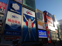 A vibrant urban scene featuring large, colorful billboards in a bustling cityscape. The central focus is a giant advertisement depicting a running man, surrounded by other illuminated signs showcasing various brands and products. The sky is clear, and the light suggests it is late afternoon.