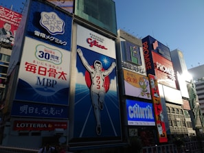 A vibrant urban scene featuring large, colorful billboards in a bustling cityscape. The central focus is a giant advertisement depicting a running man, surrounded by other illuminated signs showcasing various brands and products. The sky is clear, and the light suggests it is late afternoon.