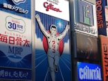 A large billboard featuring an illustration of a runner with arms raised in victory, set against a blue background with white rays emanating from the center. The surrounding area includes various advertisements and signs containing Japanese text, some with corporate logos.